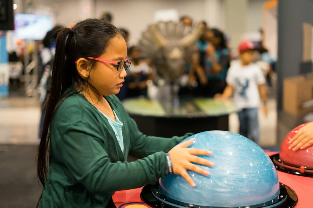 A young girl wearing glasses, touching an interactive exhibit at a science museum for children. The exhibit itself is a clear hemispherical dome bolted to a tabletop, and apparently containing some swirling, metallic blue fluid.